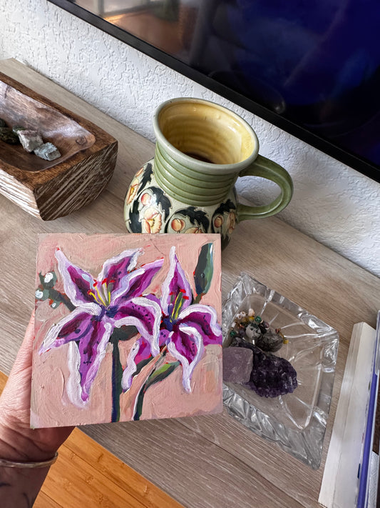 Small painting of a purple flower on a wooden block with a ceramic mug and other items in the background.