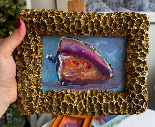 Hand holding a small abstract painting in a textured gold frame with a blurred indoor background.