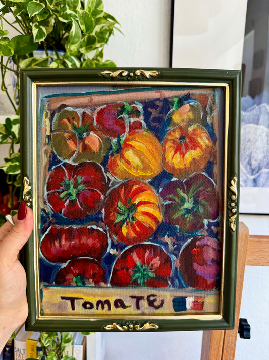 Framed painting of tomatoes with a hand holding it, set against a blurred indoor background.