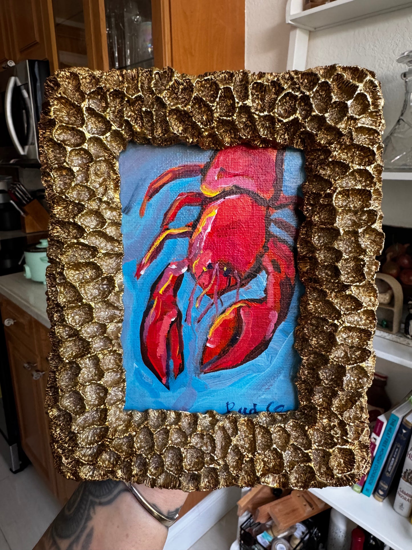 Painting of a red lobster in a textured gold frame on a blue background.