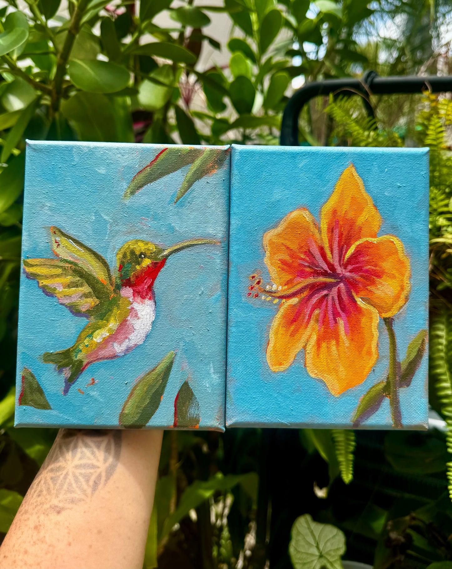Two small paintings of a hummingbird and a flower held against a natural background.
