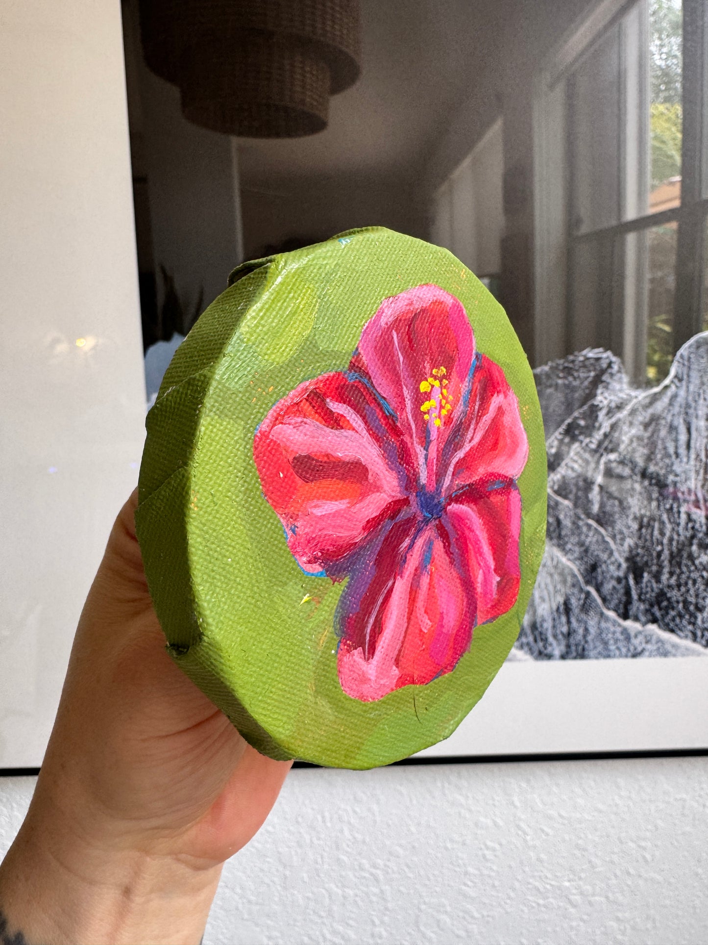A hand holds a small round canvas painting featuring a bright pink hibiscus flower on a lime green background, with a blurred indoor room and framed artwork behind it.