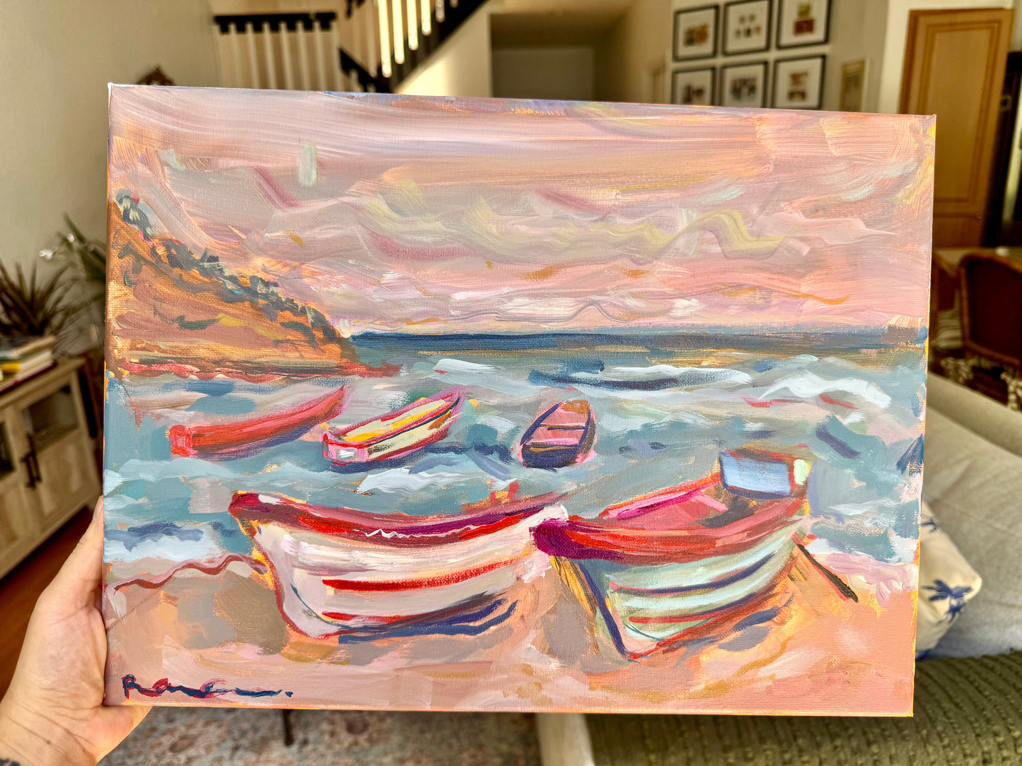 A hand holds a square canvas painting of small colorful boats on a beach at sunset. The artwork features pink, peach, blue, and red tones, with the person holding the canvas visible at the lower left in an indoor room setting.