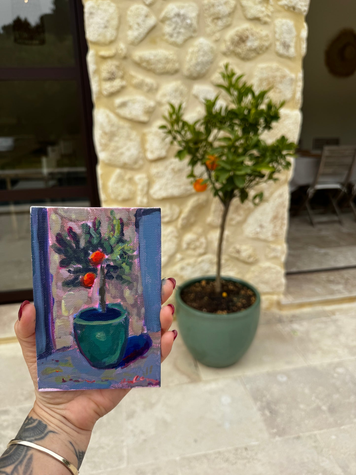 Hand holding a small painting of a green pot with plants against a textured white wall.