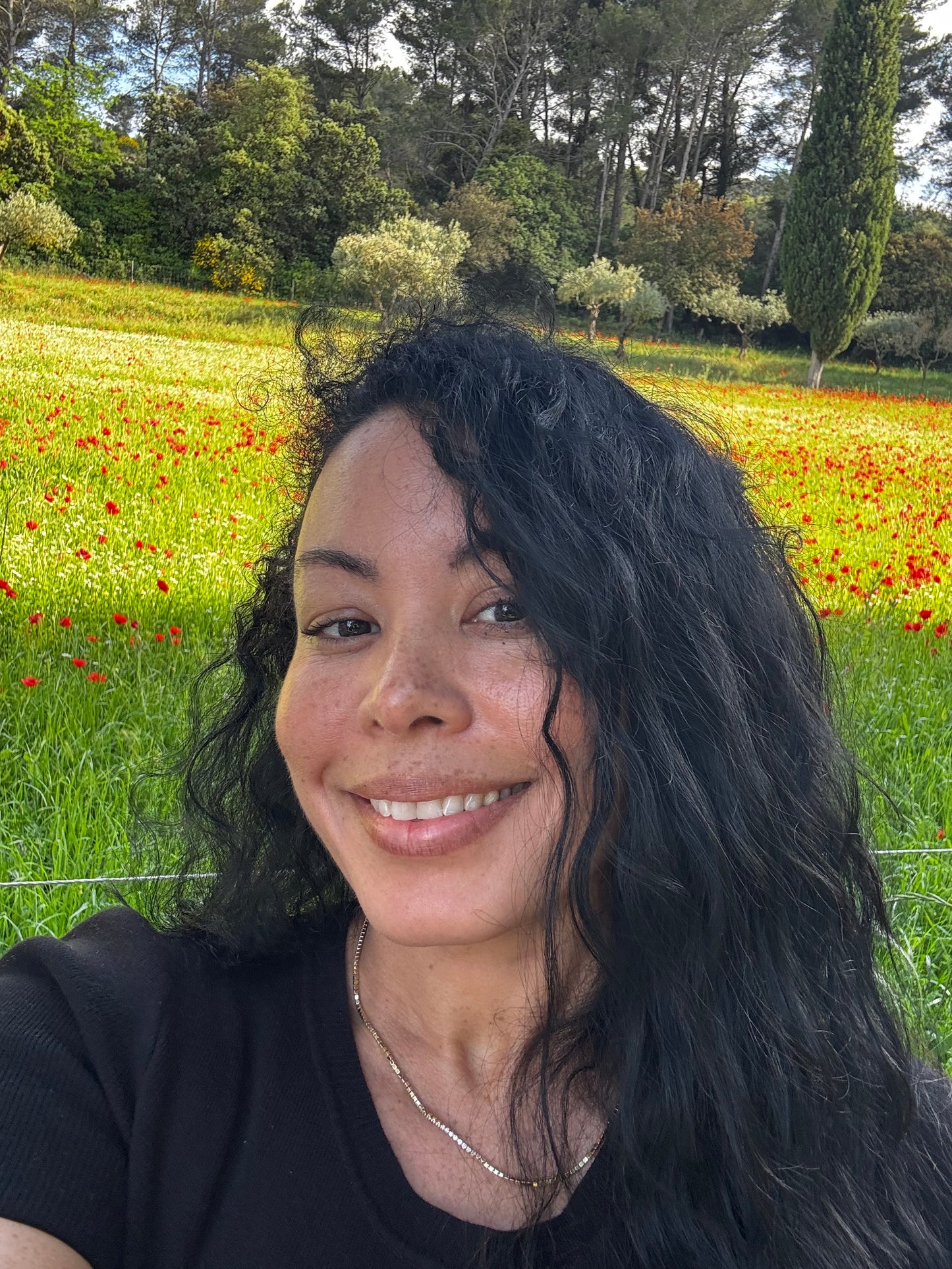 Person with long dark hair in a natural setting with greenery and flowers.