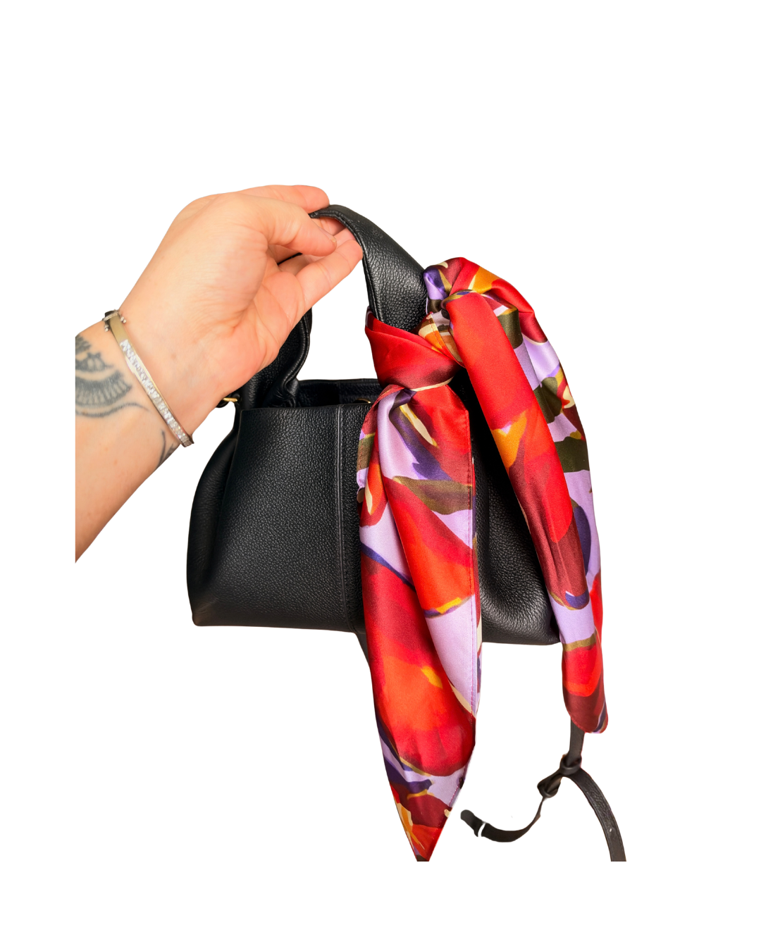 A hand wearing a silver bracelet holds a black leather handbag with a vibrant red, orange, and purple floral silk scarf tied around its handle, set against a plain white background.