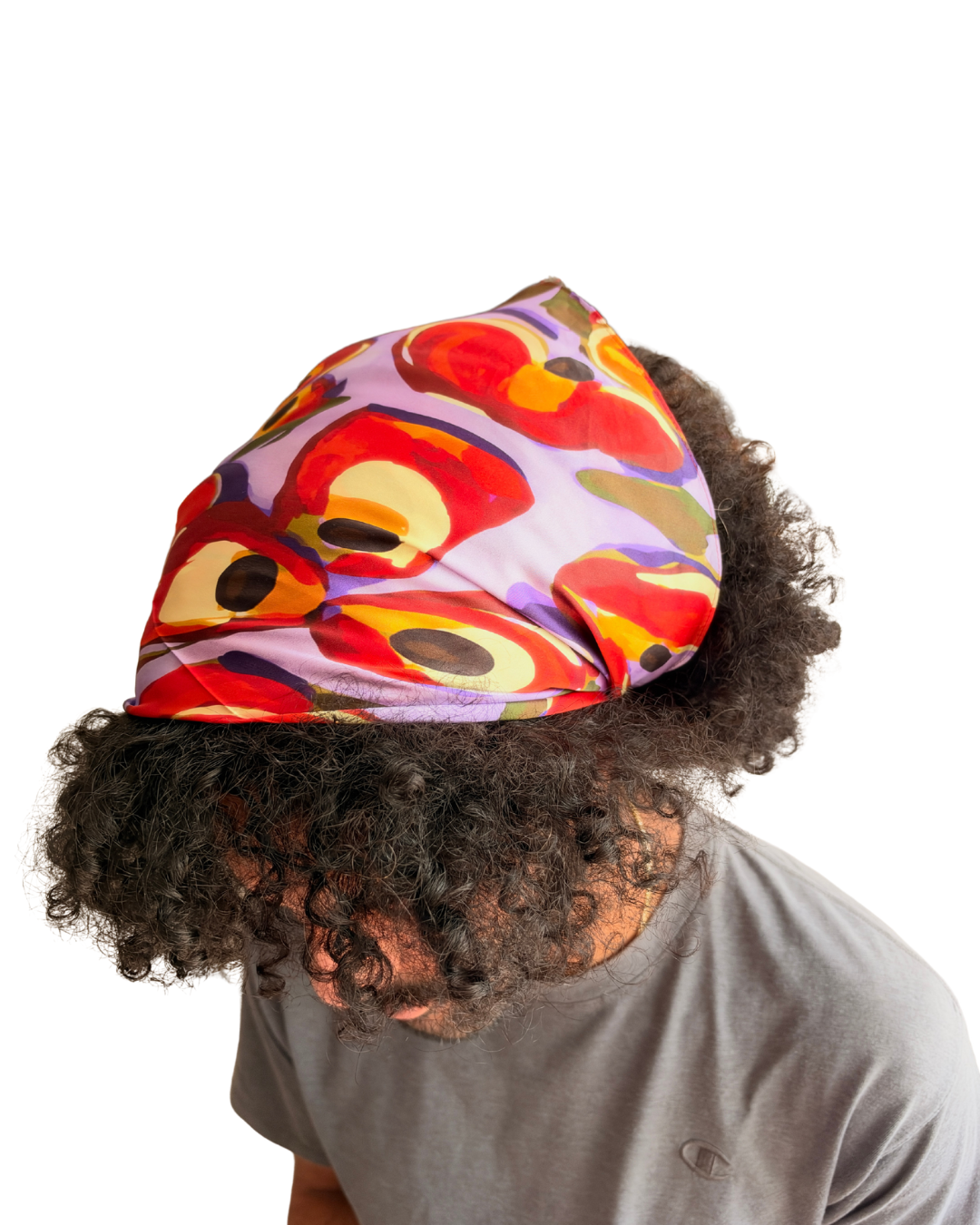 A person with curly dark hair wearing a colorful abstract-patterned headscarf and a gray t-shirt, shown from a side/back angle against a plain white background.
