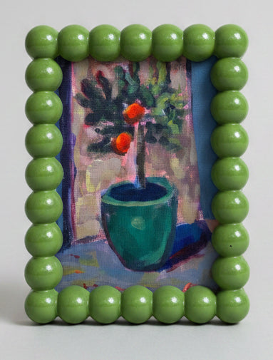 Hand holding a small painting framed with green beads, with a plant and artwork in the background.