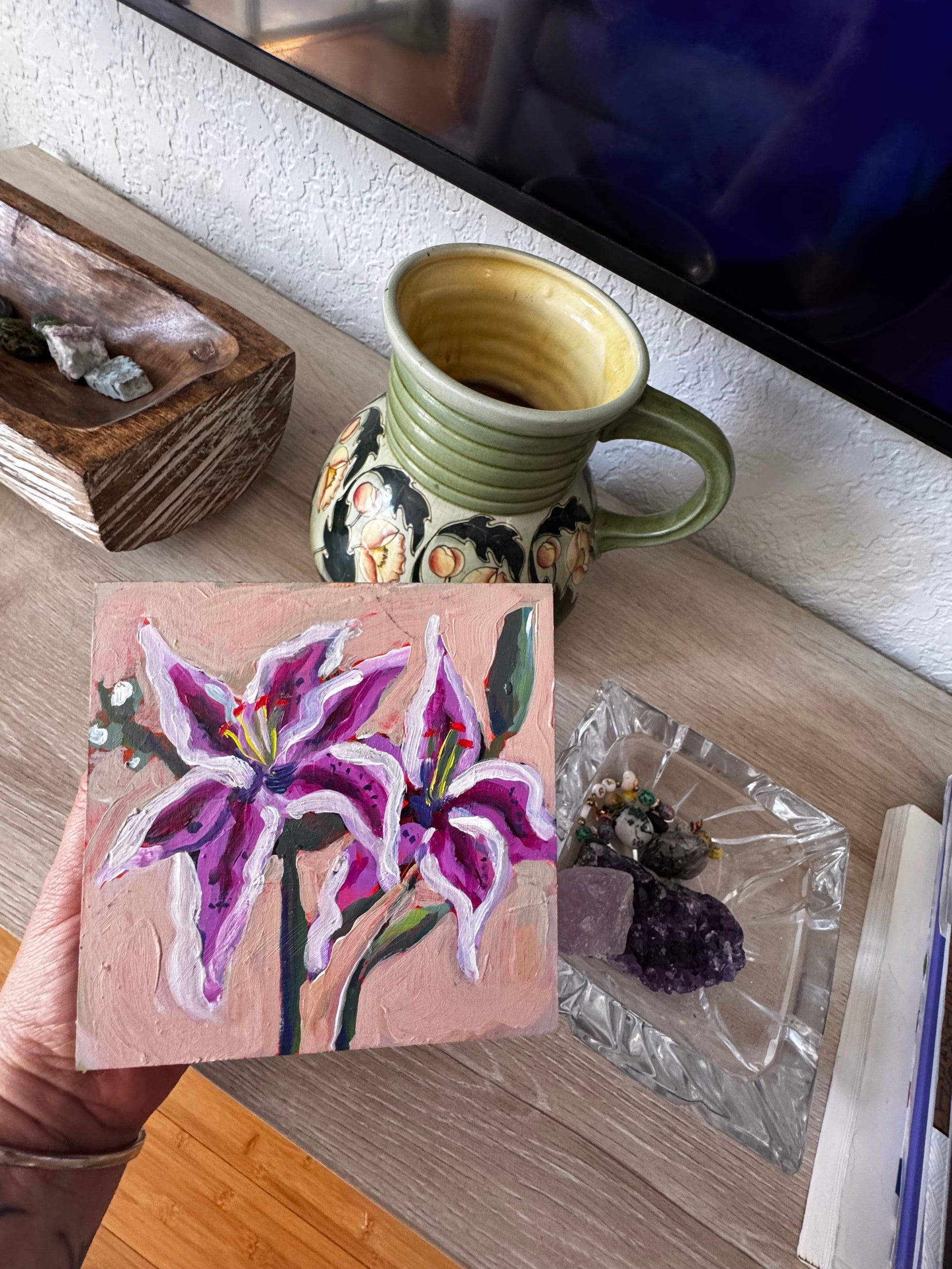 Small painting of a purple flower on a wooden block with a ceramic mug and other items in the background.