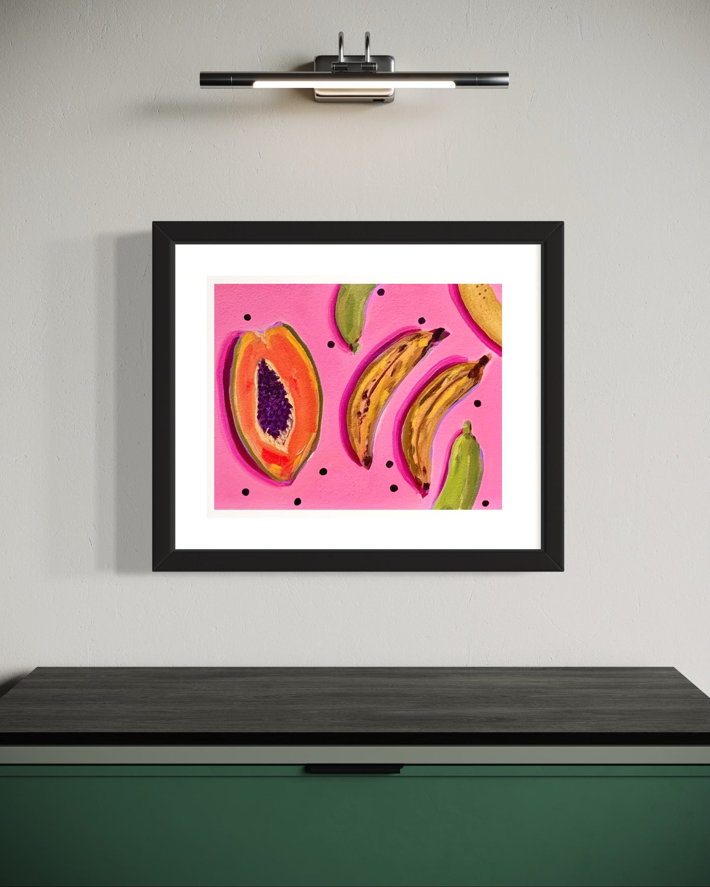 Framed artwork of tropical fruits on a pink background hanging on a wall.