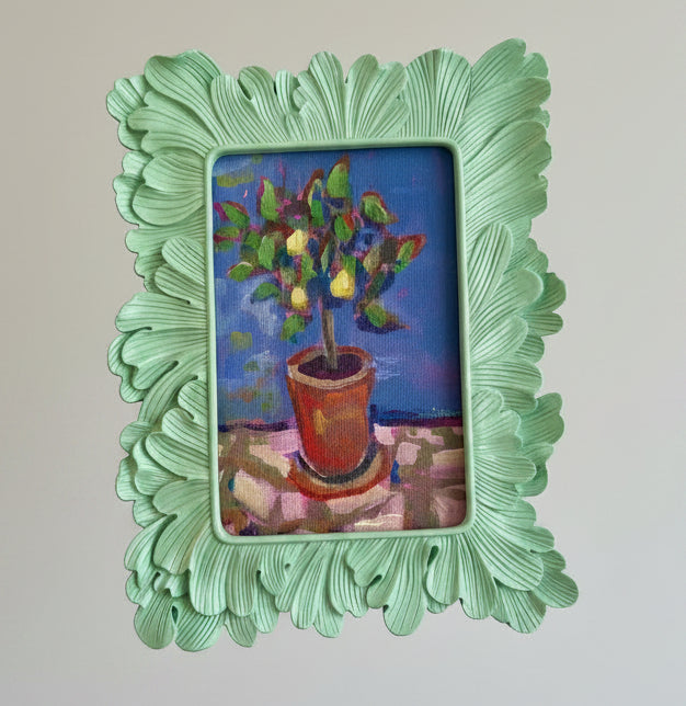Small painting of a potted plant in a decorative green frame held by a hand.