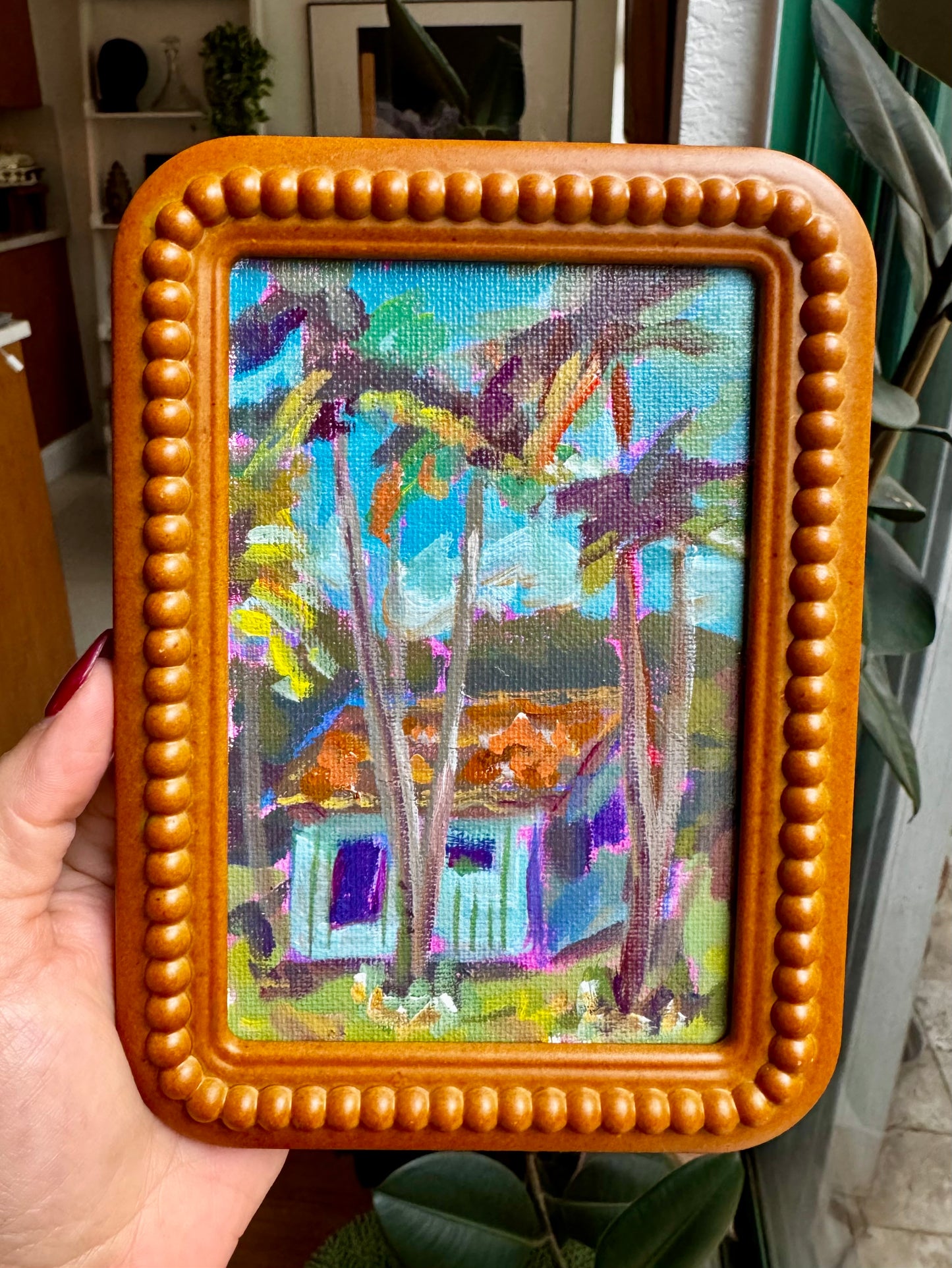 Framed painting of a tropical scene with palm trees and a house.