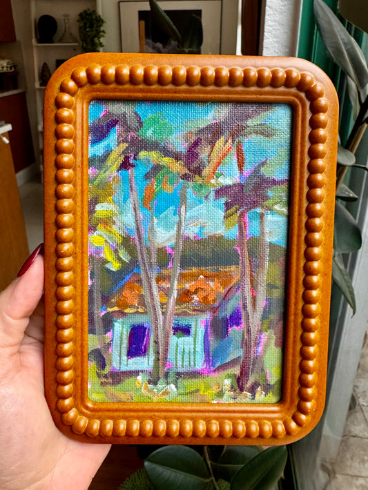 Framed painting of a tropical scene with palm trees and a house.