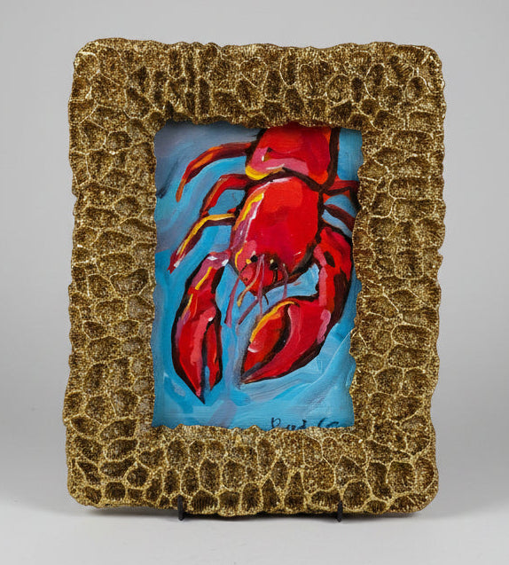 Painting of a red lobster in a textured gold frame on a white surface.
