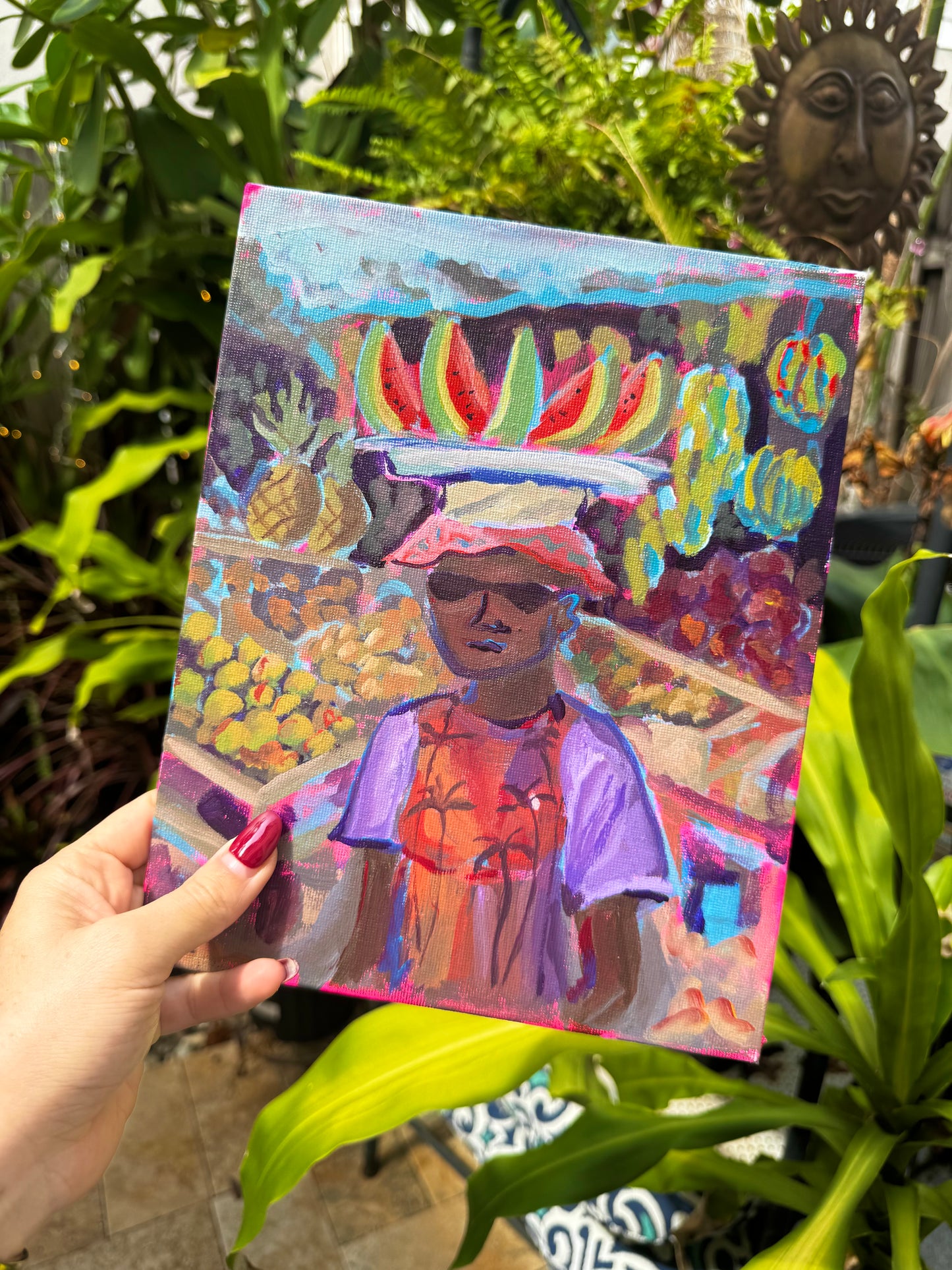 Hand holding a colorful painting of a person with fruit on their head in a garden setting