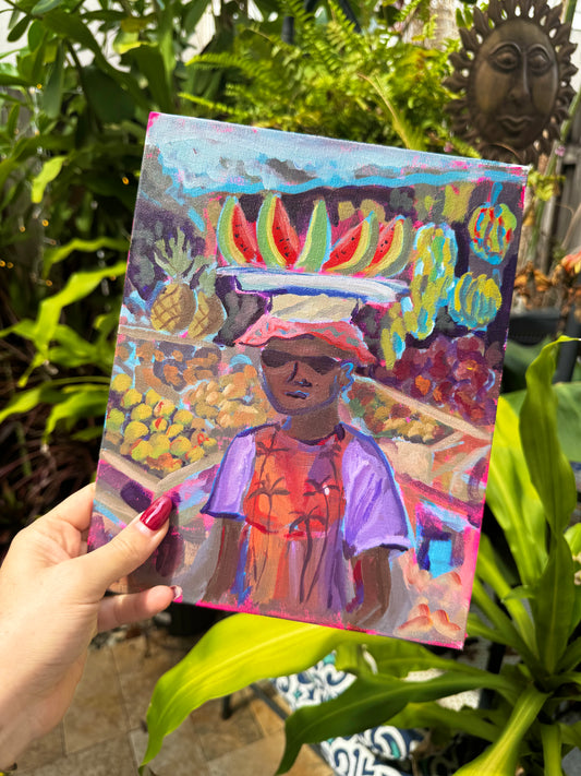 Hand holding a colorful painting of a person with fruit on their head in a garden setting