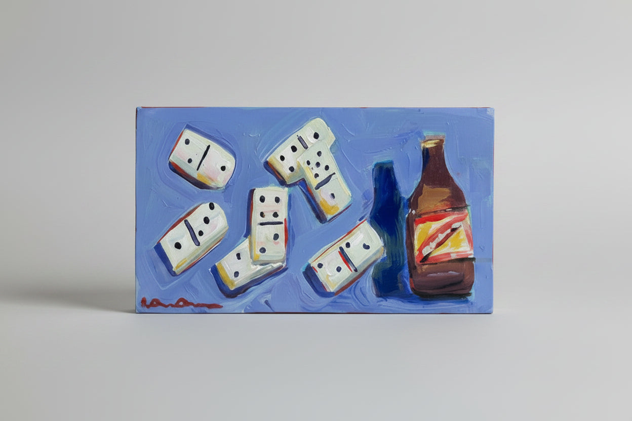A hand holds a rectangular blue painting featuring white dominoes and a brown bottle, photographed in a real indoor room with shelves, artwork, and window blinds visible in the background.