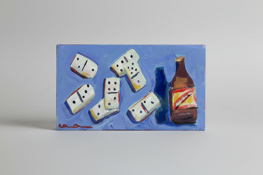 A hand holds a rectangular blue painting featuring white dominoes and a brown bottle, photographed in a real indoor room with shelves, artwork, and window blinds visible in the background.