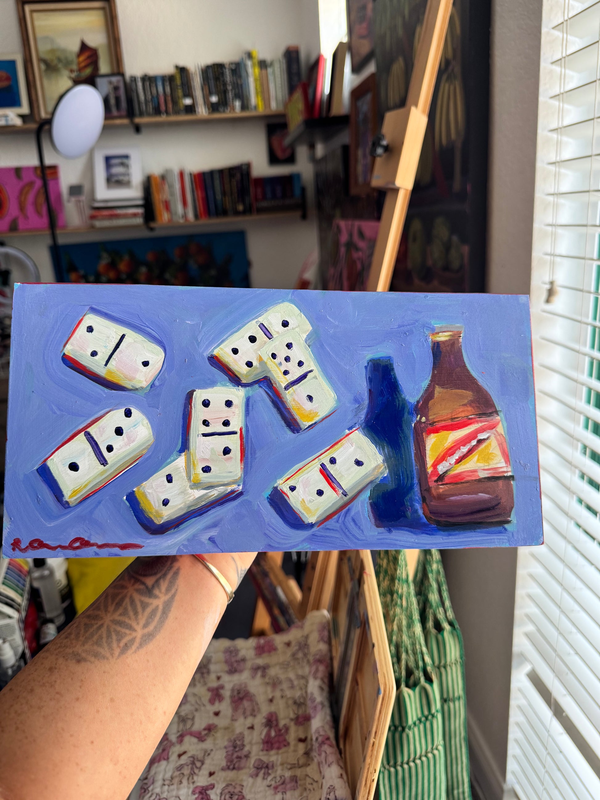 A hand holds a small painted canvas featuring white domino tiles and a brown bottle on a blue background, with an art studio and easel visible in the room behind it.