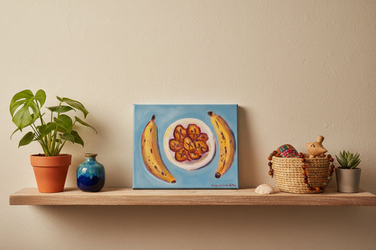 A hand holds a square canvas painting featuring two yellow bananas on either side of a plate of sliced bananas, set against a light blue background, with a wooden fence and string lights behind it.