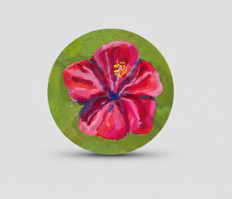 A hand holds a round canvas painting featuring a vibrant pink hibiscus flower with yellow and blue details on a textured green background.