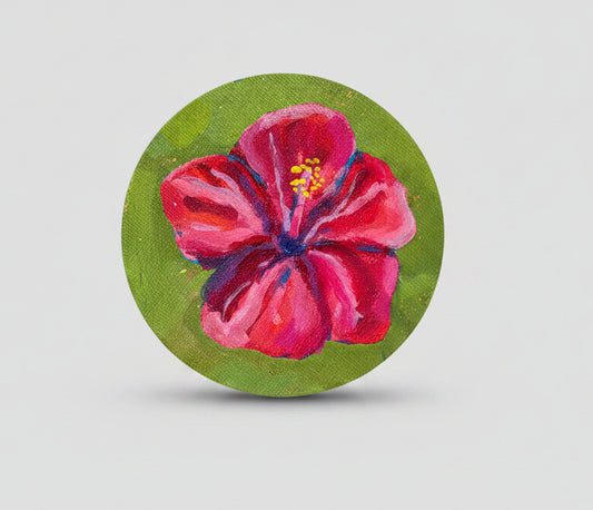 A hand holds a round canvas painting featuring a vibrant pink hibiscus flower with yellow and blue details on a textured green background.