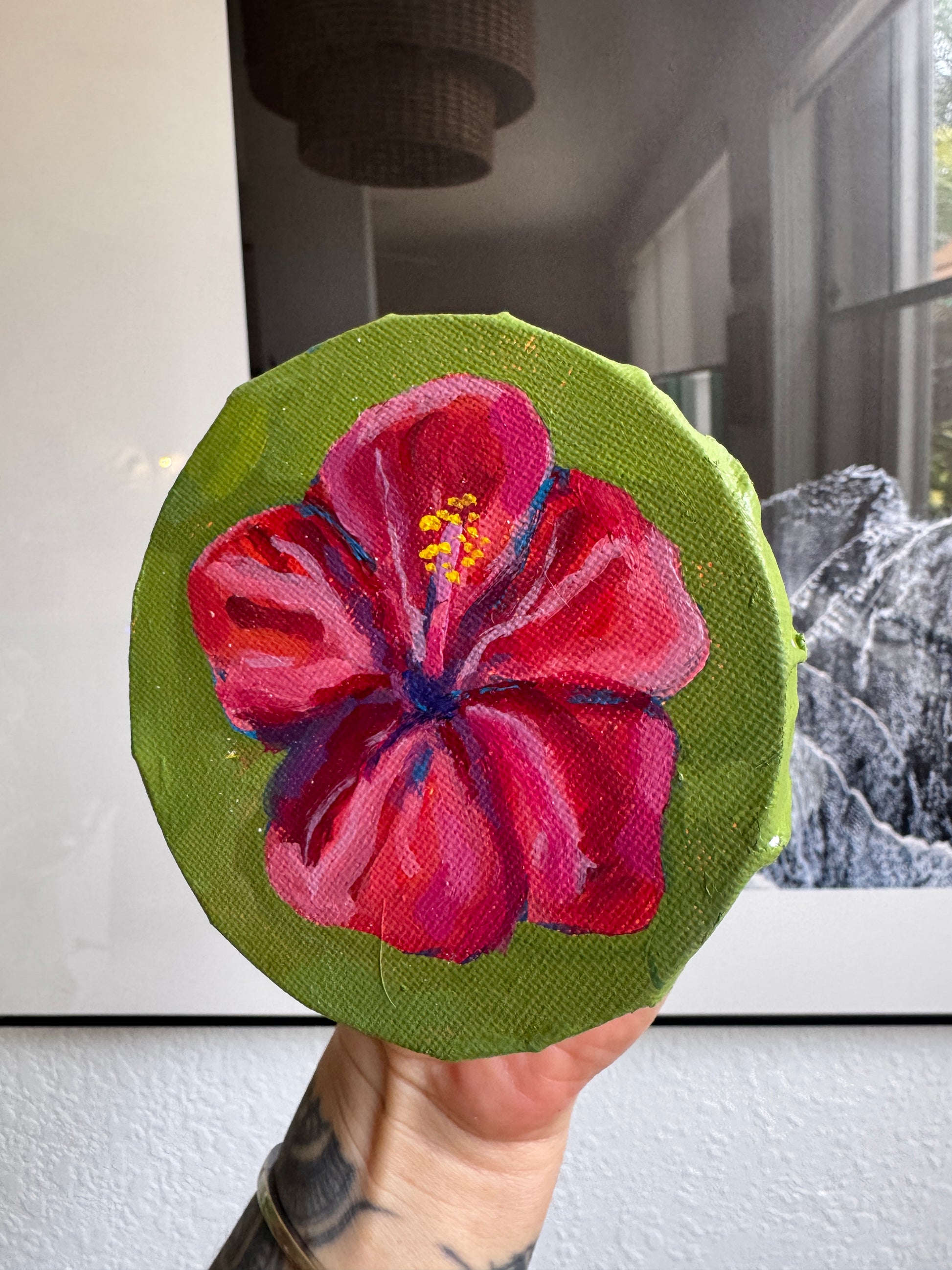 A hand holds a round canvas painting featuring a vibrant pink hibiscus flower on a green background, with a blurred indoor environment behind it.