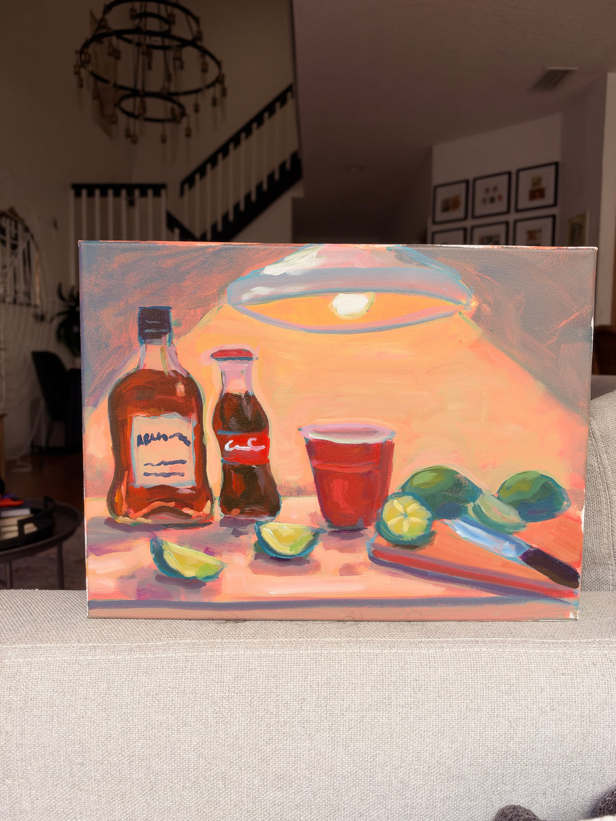 A colorful acrylic painting of a still life scene featuring liquor bottles, a glass of red drink, limes, and a cutting board, displayed on a light-colored sofa in a living room setting.