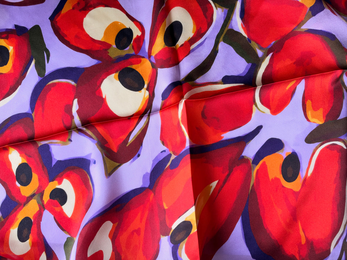A close-up of a patterned fabric featuring large, stylized red flowers with cream centers and dark outlines on a light purple background.