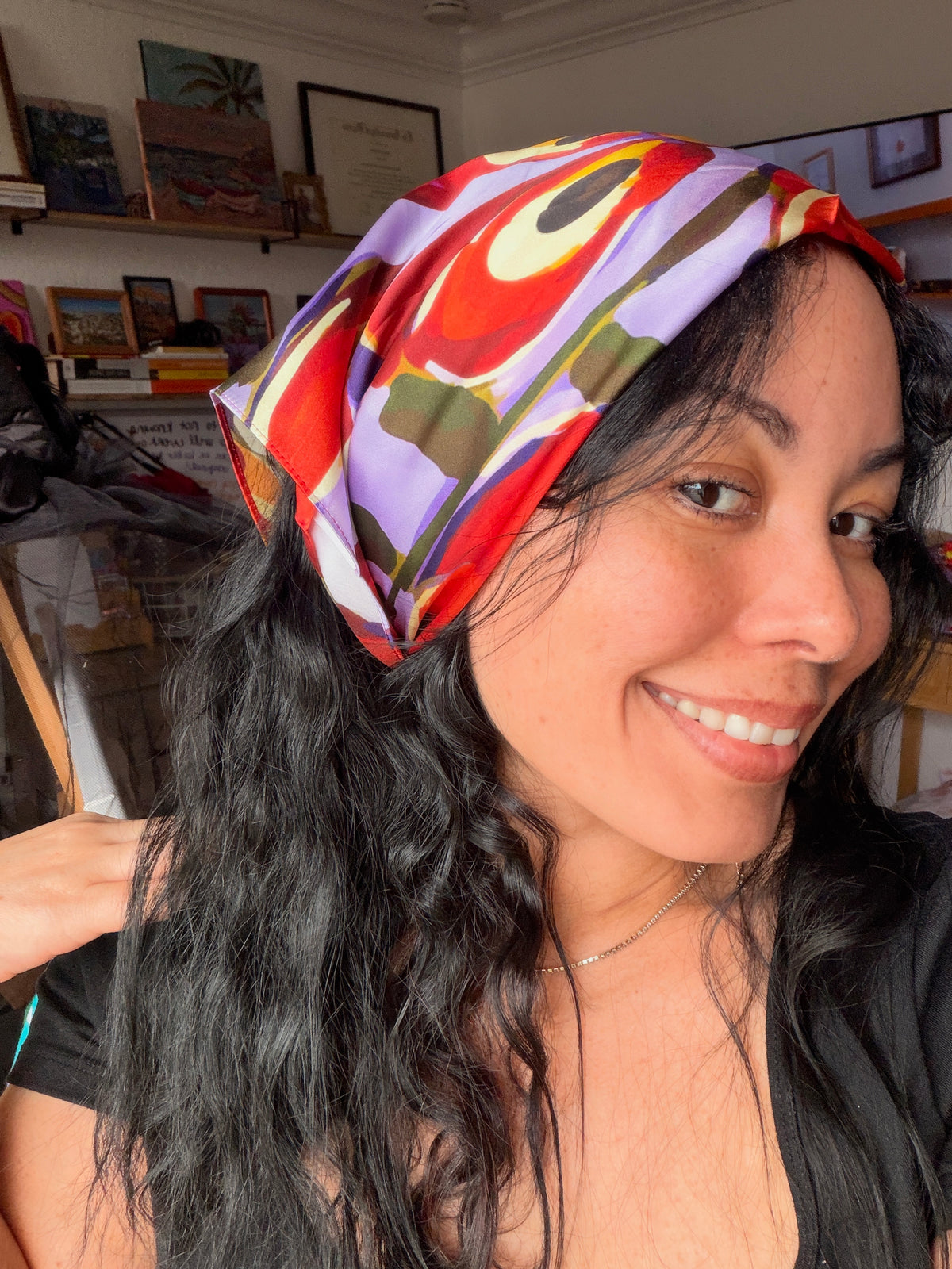 A woman wearing a colorful abstract-patterned silk headscarf in red, purple, green, and yellow, photographed in a home studio environment.