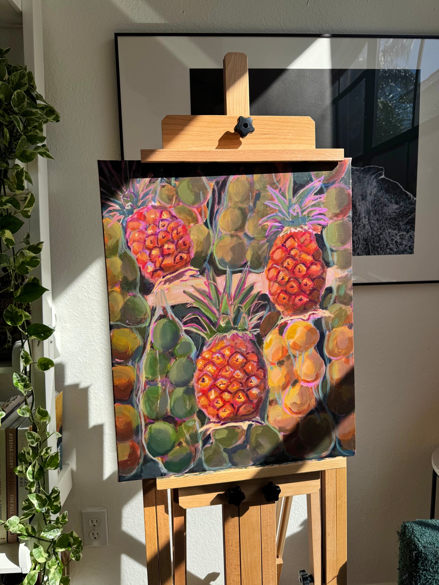Painting of pineapples on an easel in a room with plants and a TV.