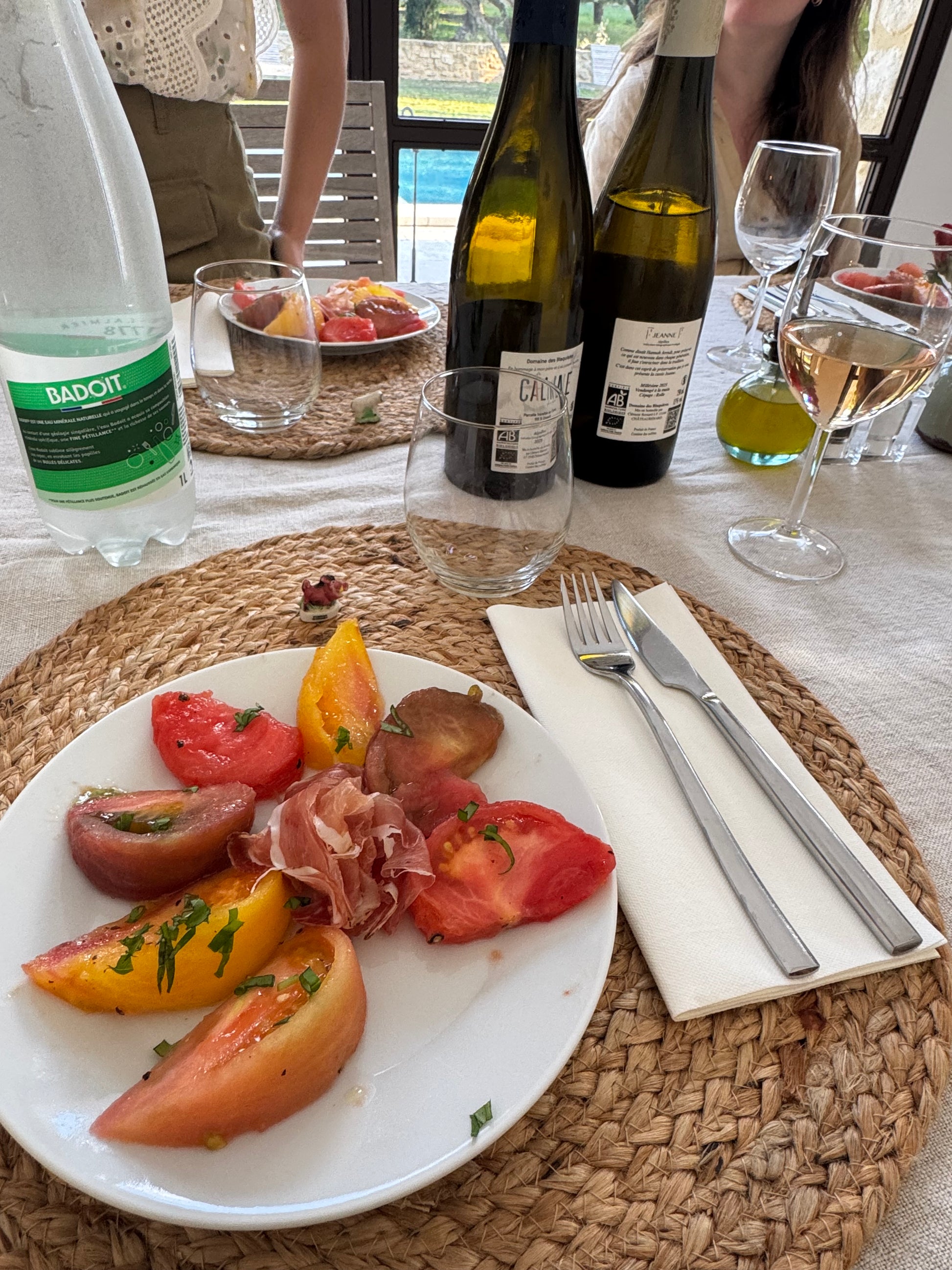 Table setting with wine bottles, glasses, and a plate of food on a woven placemat.