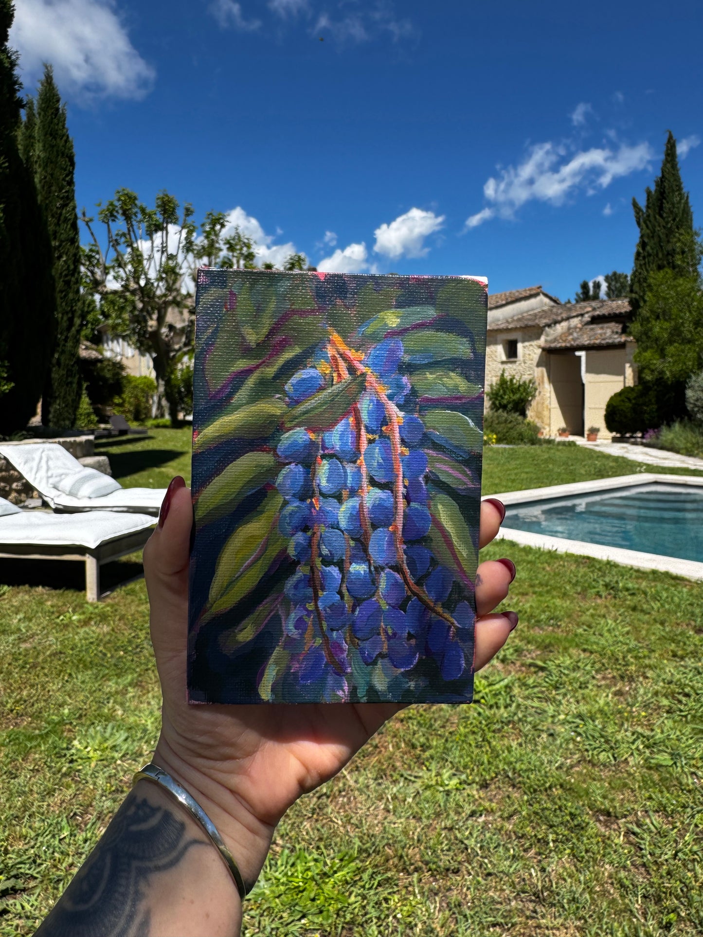Hand holding a small painting of blue flowers against a garden background with a pool and house.