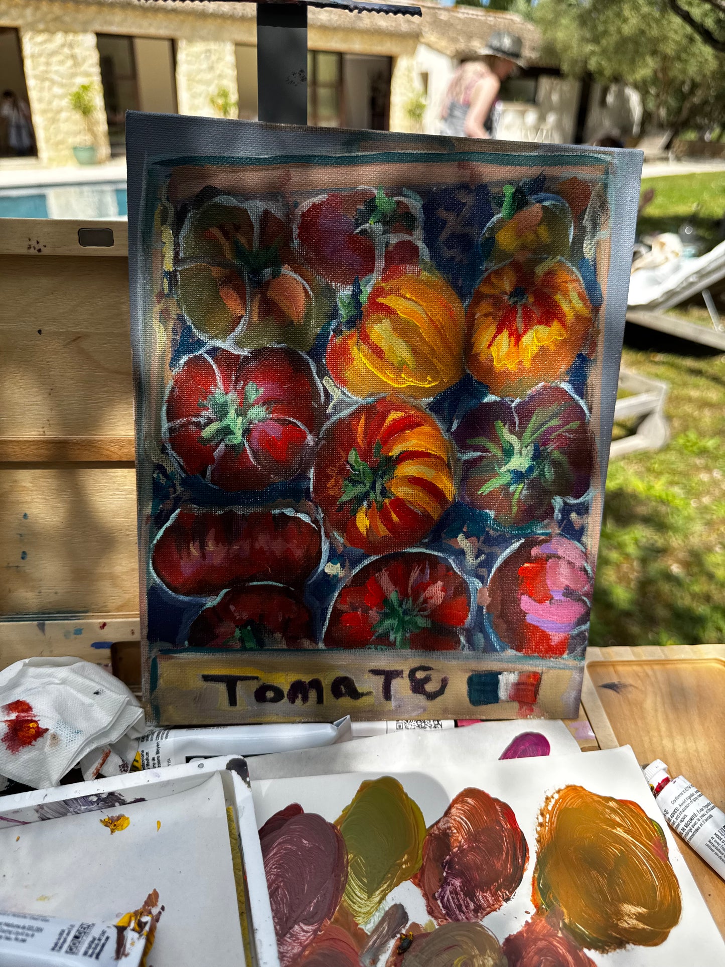 Painting of tomatoes on a canvas with a palette and paint tubes in the background.