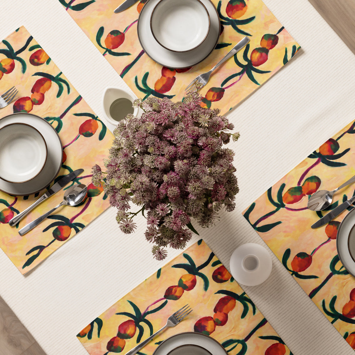 A top-down view of a styled dining table setting featuring four matching placemats with a vibrant orange and green botanical pattern on a warm cream background. The placemats are arranged around a central bouquet of dusty pink flowers, with white ceramic plates, silver cutlery, and a white tablecloth visible.