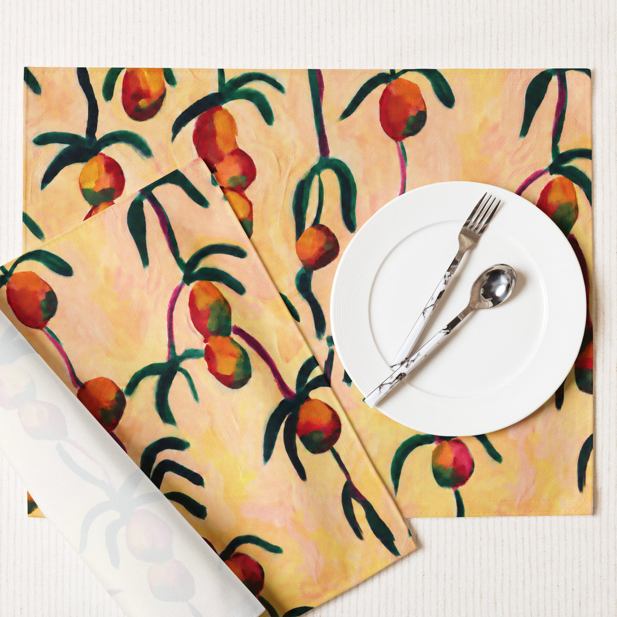 A set of table linens featuring a painted orange and green fruit pattern on a warm yellow background, styled with a white plate and silver cutlery on a white textured tabletop.