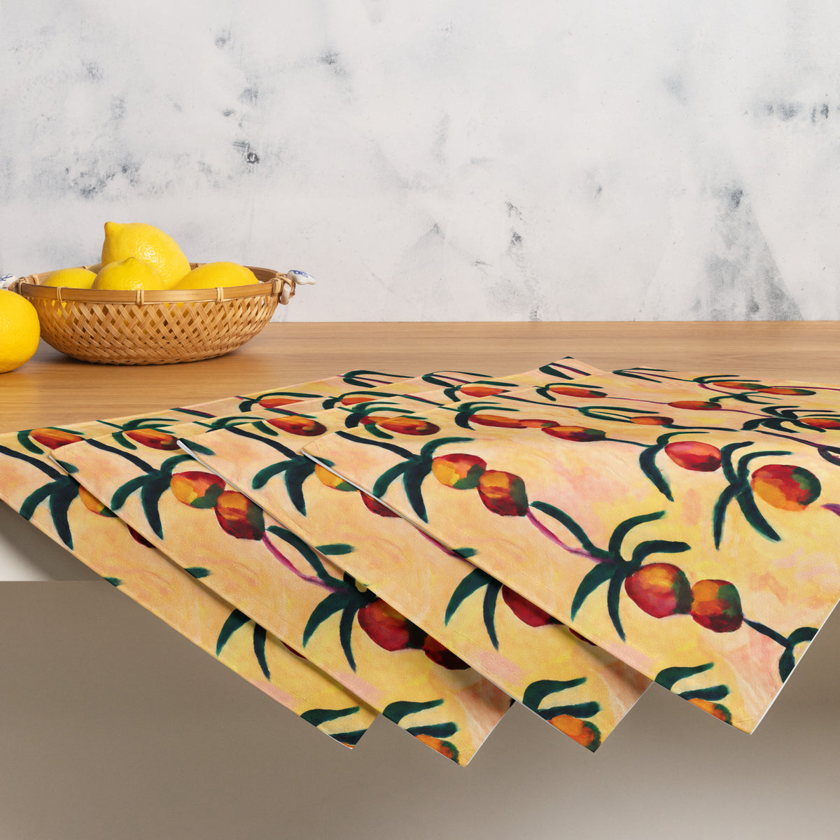 Three rectangular placemats with a colorful fruit branch pattern are fanned out on a light wood table. The background features a marble wall and a woven basket of lemons.